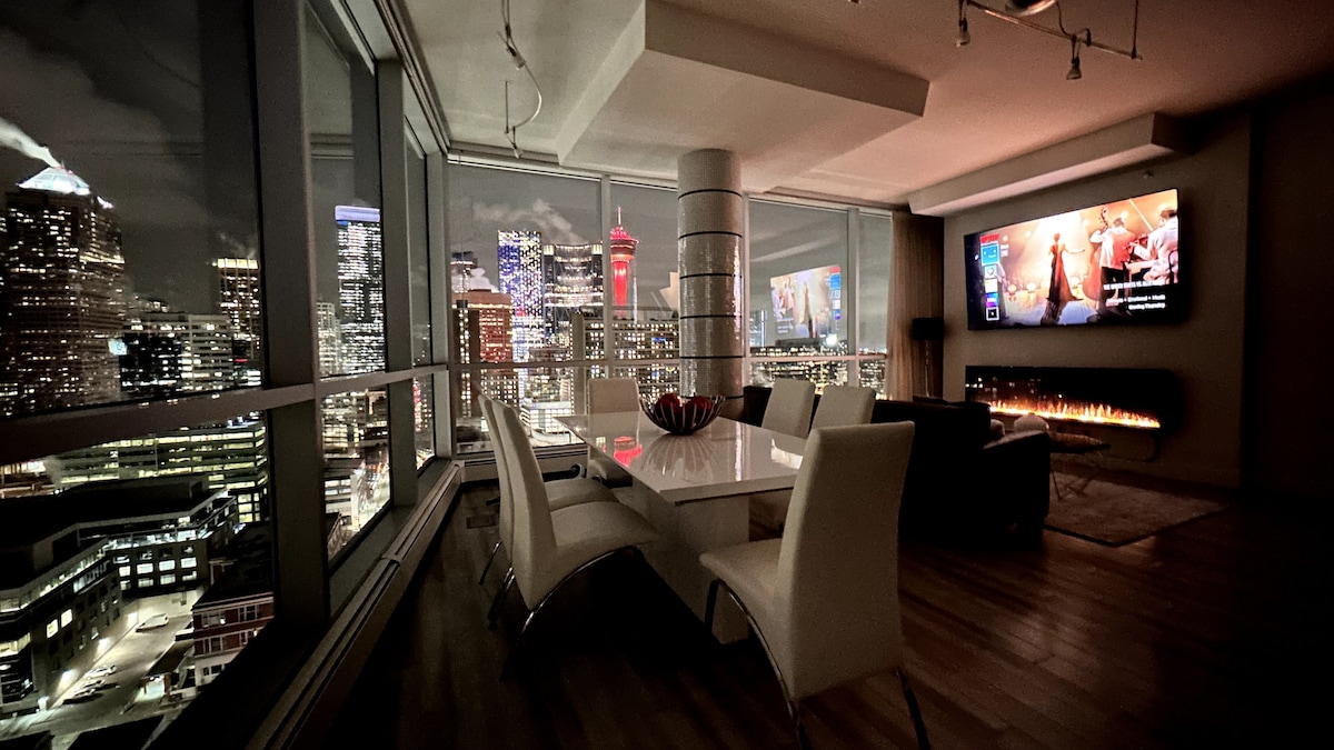 The modern dining area features a sleek white table surrounded by comfortable chairs, set against a backdrop of expansive floor-to-ceiling windows showcasing vibrant views of the Calgary skyline at night. A wall-mounted television and a cozy fireplace add to the inviting atmosphere.