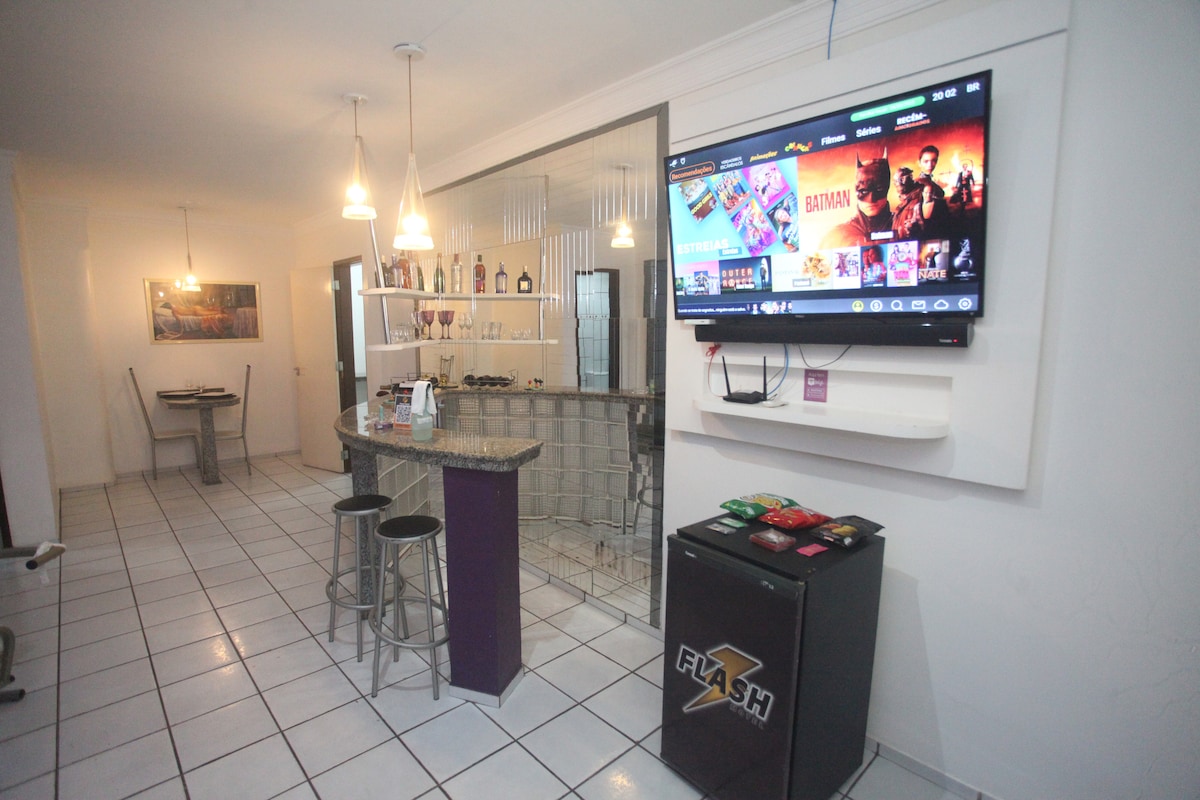 A modern living area features a large wall-mounted television displaying various entertainment options. Bar stools are positioned around a counter, with a glimpse of a dining table visible in the background. Soft lighting from pendant fixtures enhances the inviting ambiance.