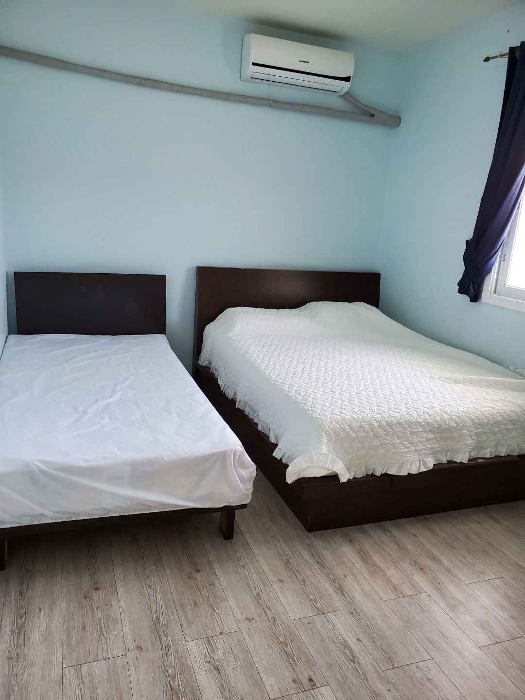 A light blue bedroom features two beds, one spacious with a white textured coverlet and one single with a white fitted sheet. An air conditioning unit is mounted on the wall, and a window with dark curtains allows natural light to enter.