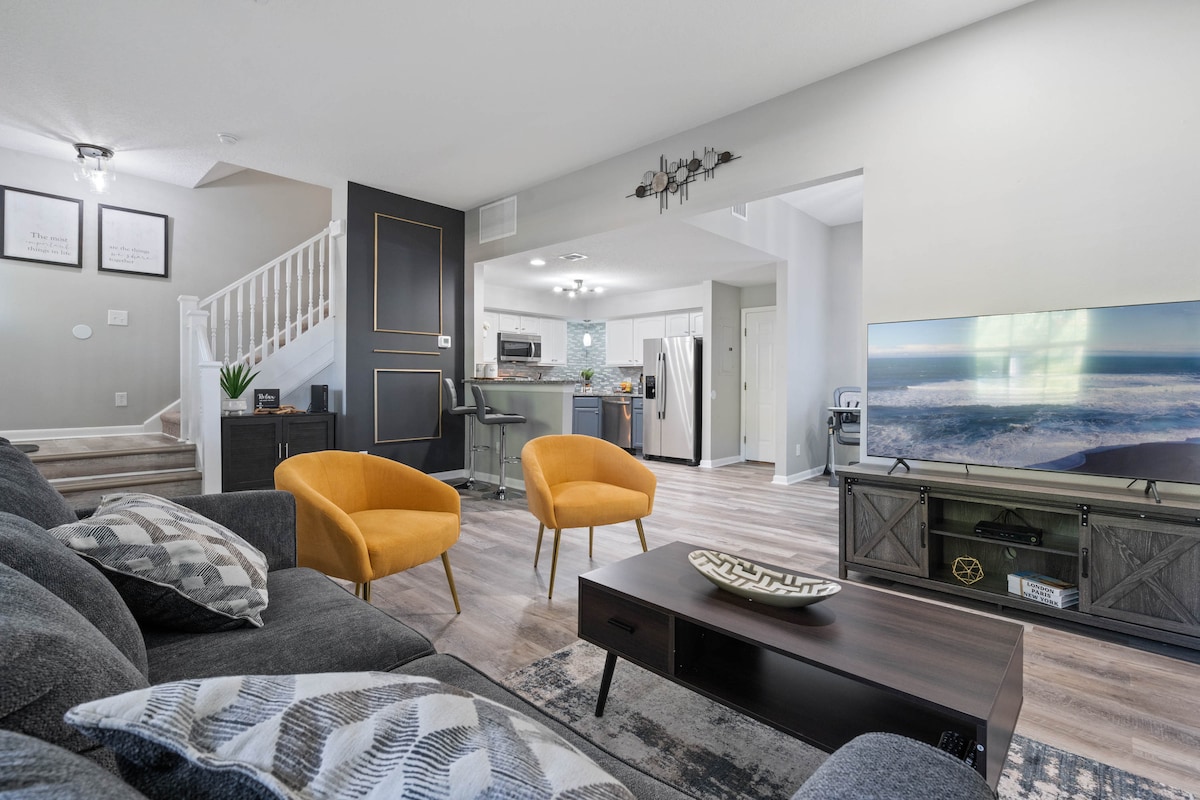 A spacious living area features a comfortable gray sofa paired with two bright yellow chairs. A coffee table sits in front, while a television is positioned on a stand across from the seating. The open layout connects to a modern kitchen, visible in the background.