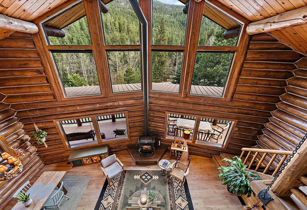 The living room is highlighted by floor-to-ceiling windows that provide views of the wooded mountainside. Natural wood walls and ceiling beams create a warm environment. Seating includes a patterned rug beneath a coffee table, surrounded by chairs and a small side table, with a wood stove positioned for warmth.