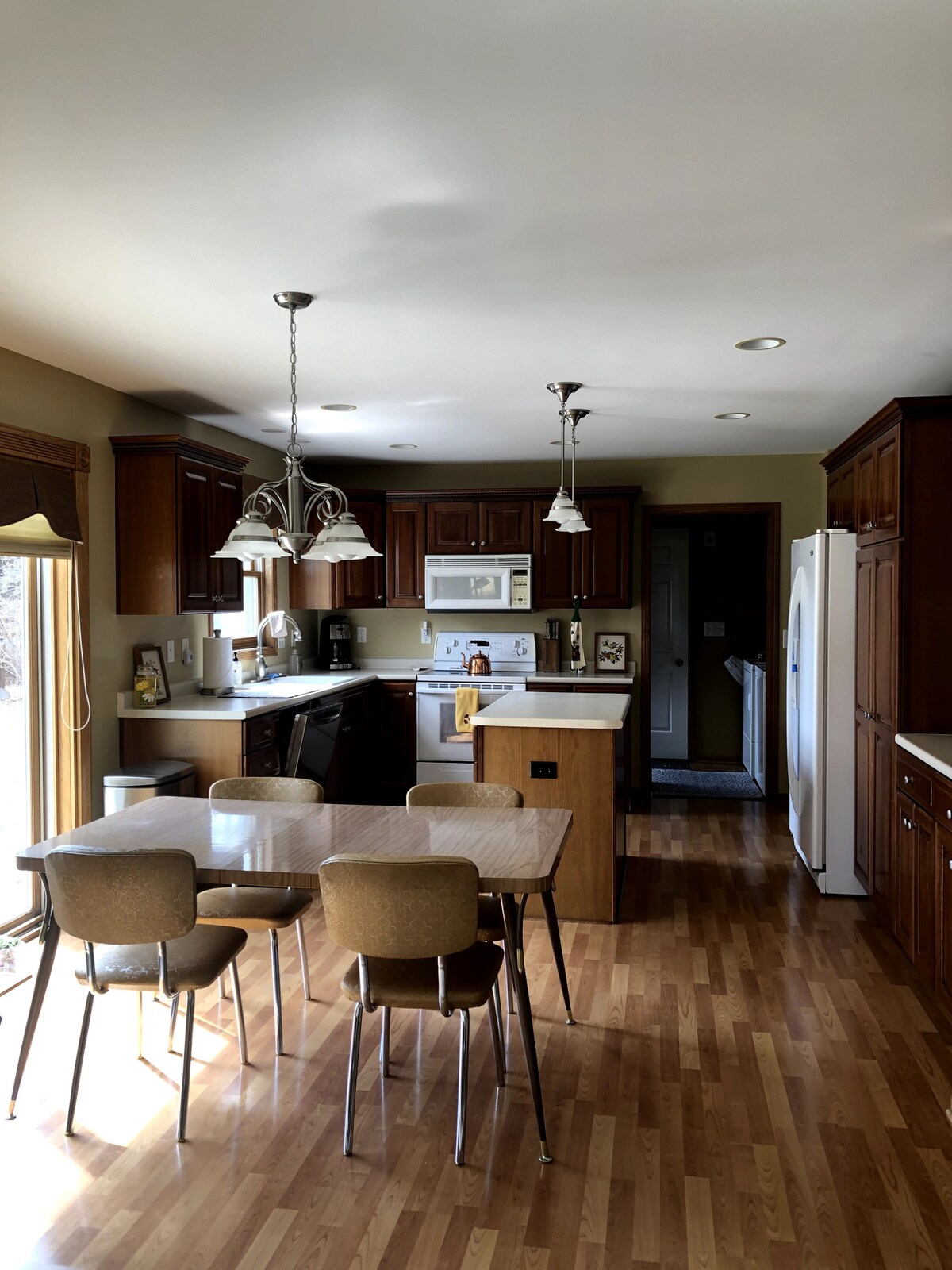 The kitchen features wooden cabinetry, white appliances, and a central island. A small dining table with four chairs is positioned beside a window, allowing natural light to enter. Additional appliances such as a coffee maker and microwave are present, along with a view into the adjoining hallway.