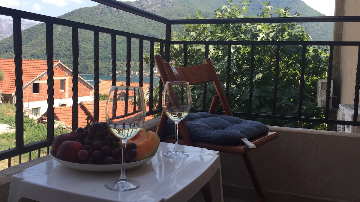 A balcony view features a small table with a plate of assorted fruits and two glasses of white wine. A wooden chair is positioned nearby, while green foliage and distant mountains are visible in the background, alongside rooftops and a glimpse of water.
