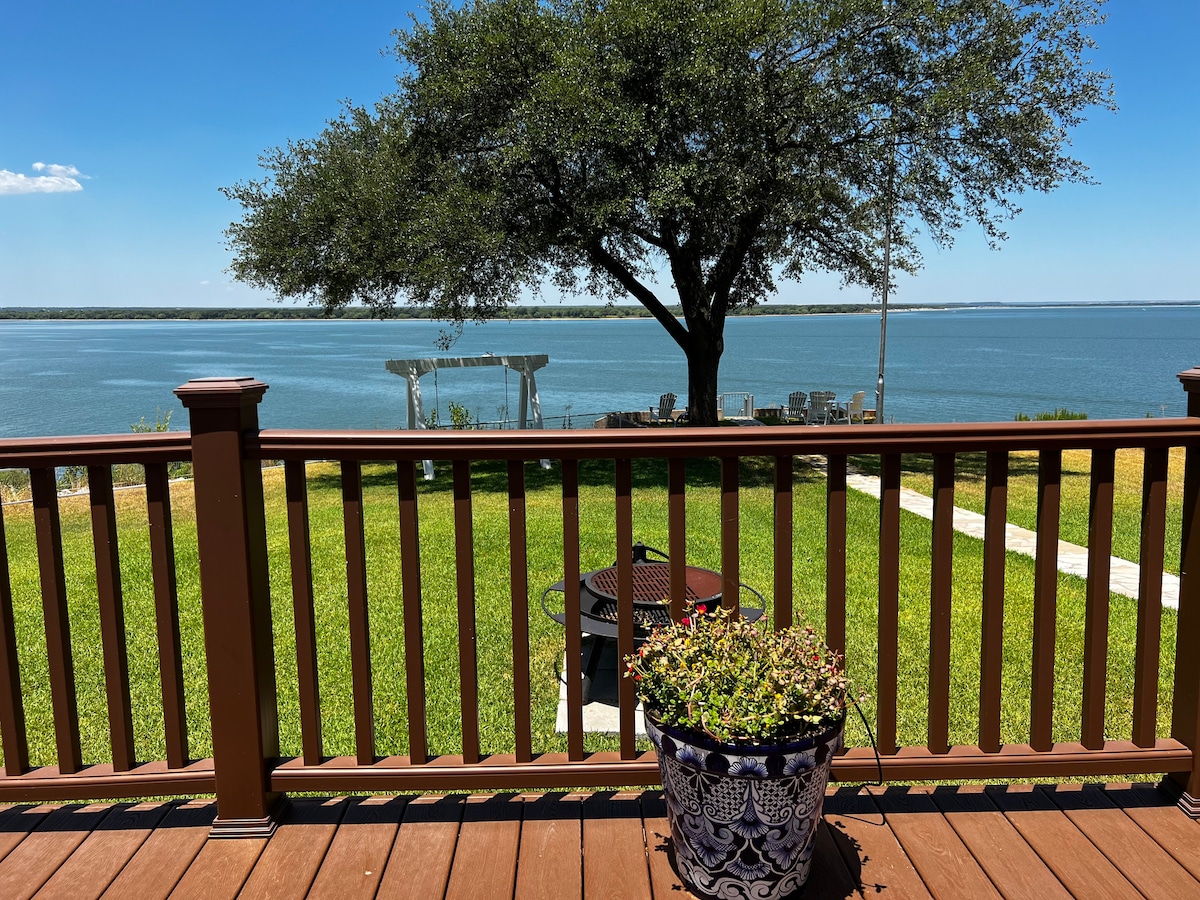 A view from the deck showcases a tranquil lake bordered by a grassy area. A large tree provides shade, while a flowerpot adds a touch of greenery. In the distance, seating areas can be seen along the water, indicating inviting outdoor spaces for relaxation.
