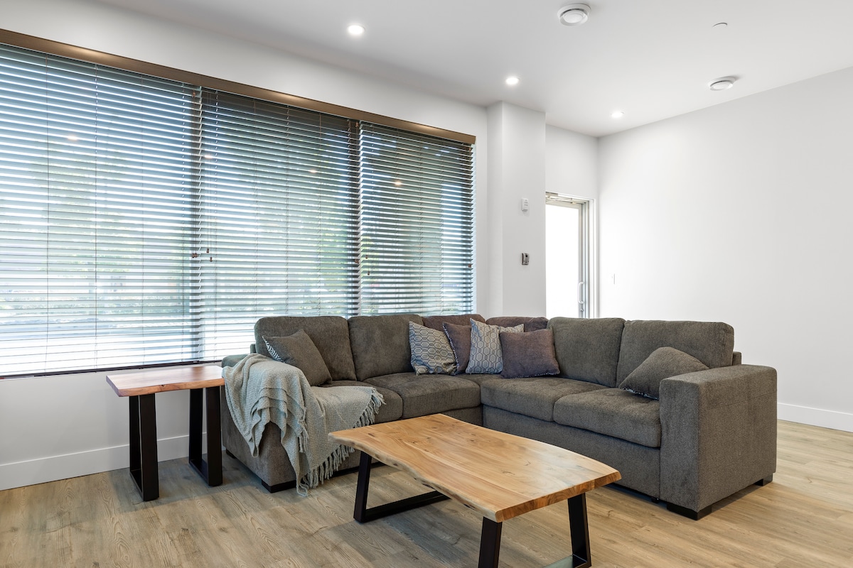 A cozy living area features a large sectional sofa adorned with decorative cushions. Natural light filters through horizontal blinds on the windows, illuminating a wooden coffee table and side table positioned on a light-colored floor.