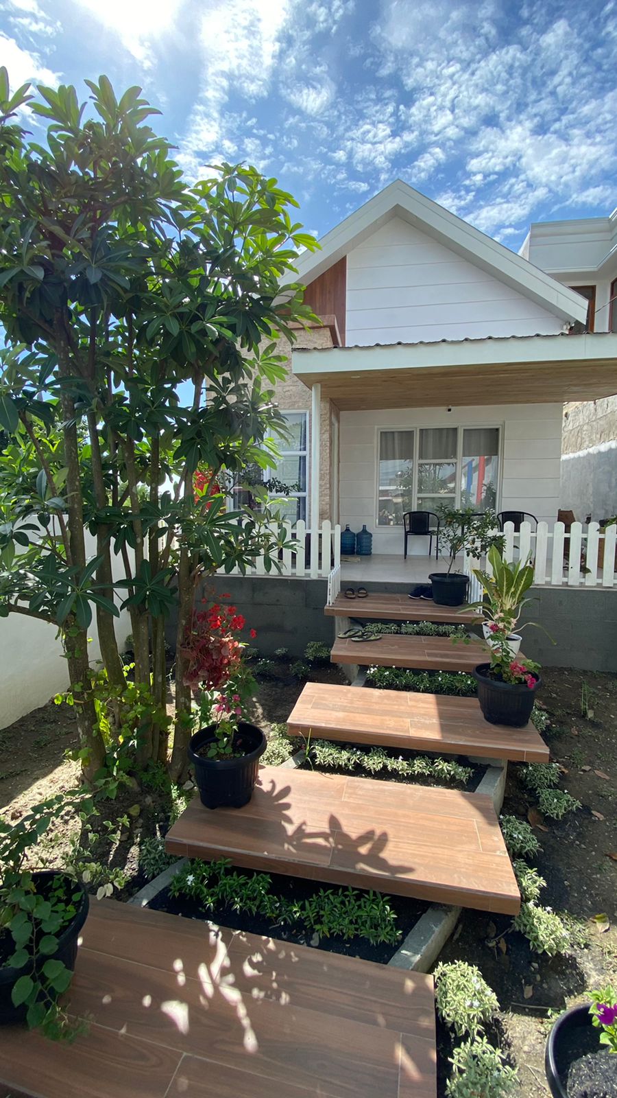 A welcoming entrance features wooden steps leading to a white-framed house, surrounded by lush greenery and potted plants. A white picket fence provides a quaint boundary, while clear blue skies enhance the inviting appearance of the homestay.