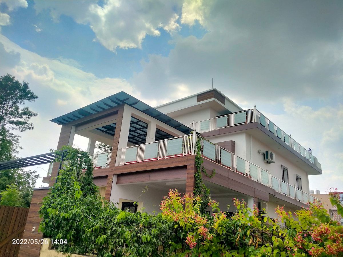 A modern villa is showcased with a clean, angular design. Multiple balconies are visible, framed by glass railings, and lush greenery surrounds the structure. The sky above features a mix of clouds, adding to the serene atmosphere.