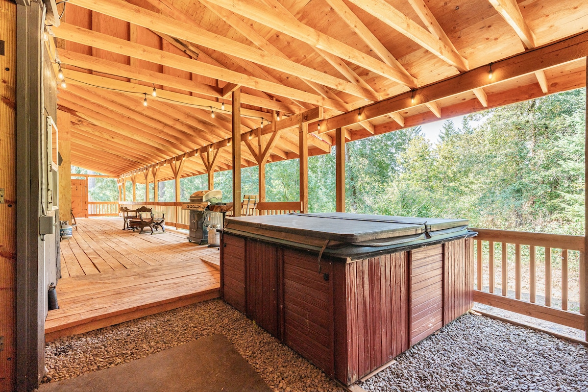 A spacious outdoor deck is covered and features wooden beams overhead. A hot tub is positioned on one side, and a seating area with a table and chairs is available for guests. The surrounding natural landscape adds to the tranquil setting.