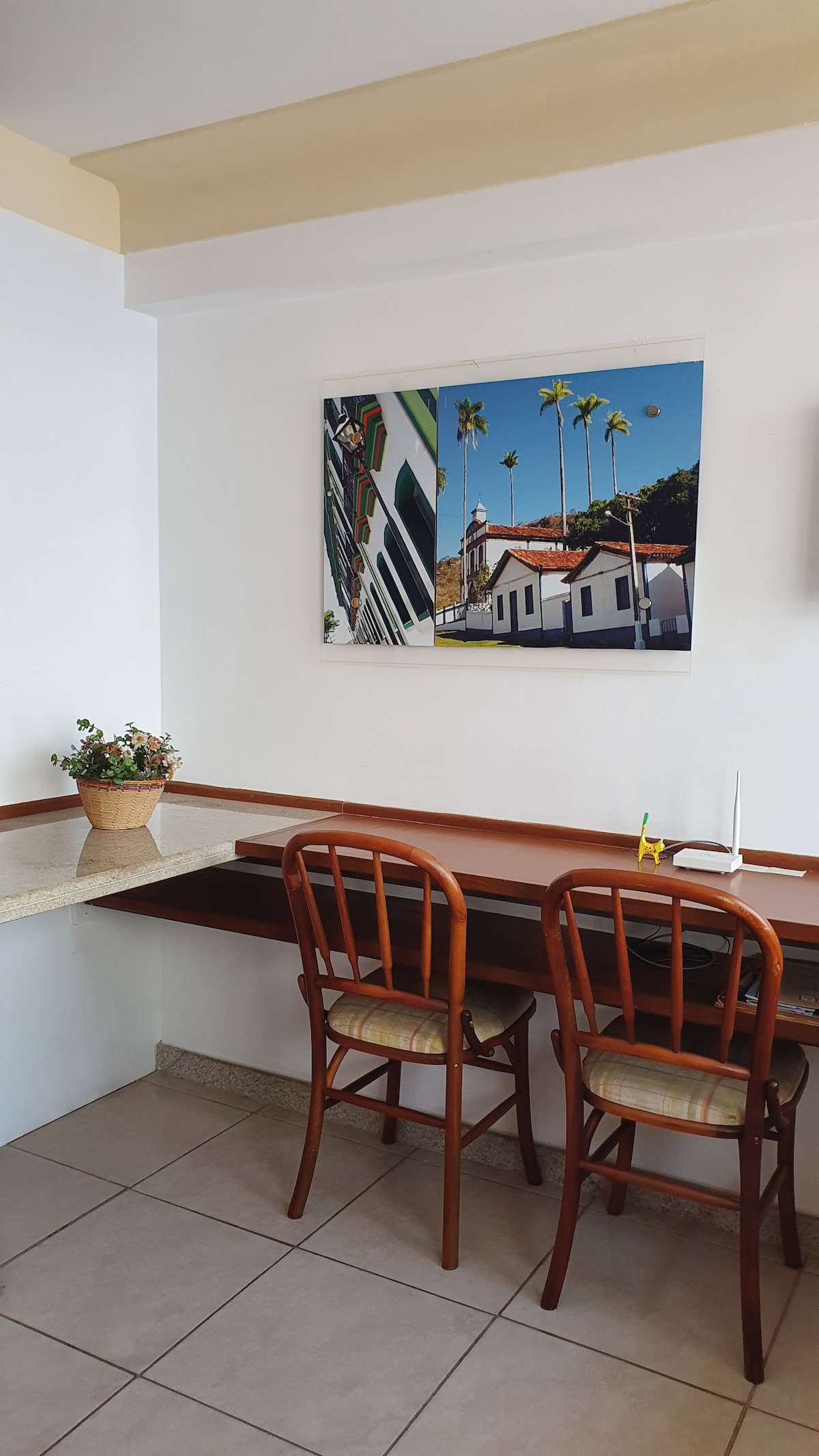 A small workspace is presented with a wooden table and two wooden chairs. A potted plant adds a touch of greenery. The wall is decorated with a vibrant photograph depicting palm trees and historical architecture.