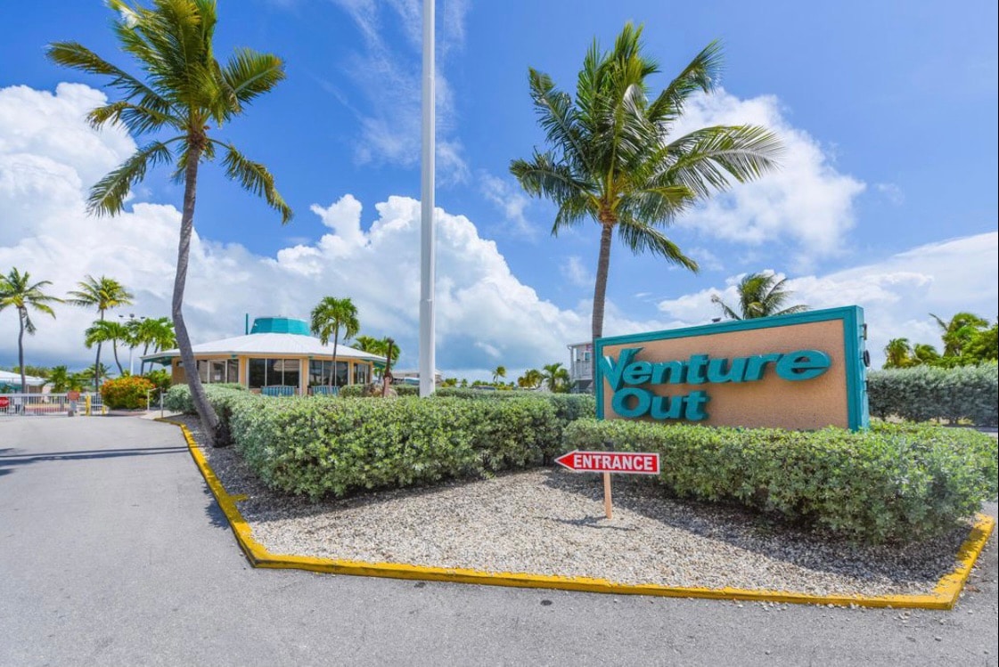 The entrance to Venture Out is marked by a prominently displayed sign amidst tropical landscaping. Lush palms sway gently, and the sky is bright with a few fluffy clouds, creating a welcoming atmosphere for guests arriving at the community.