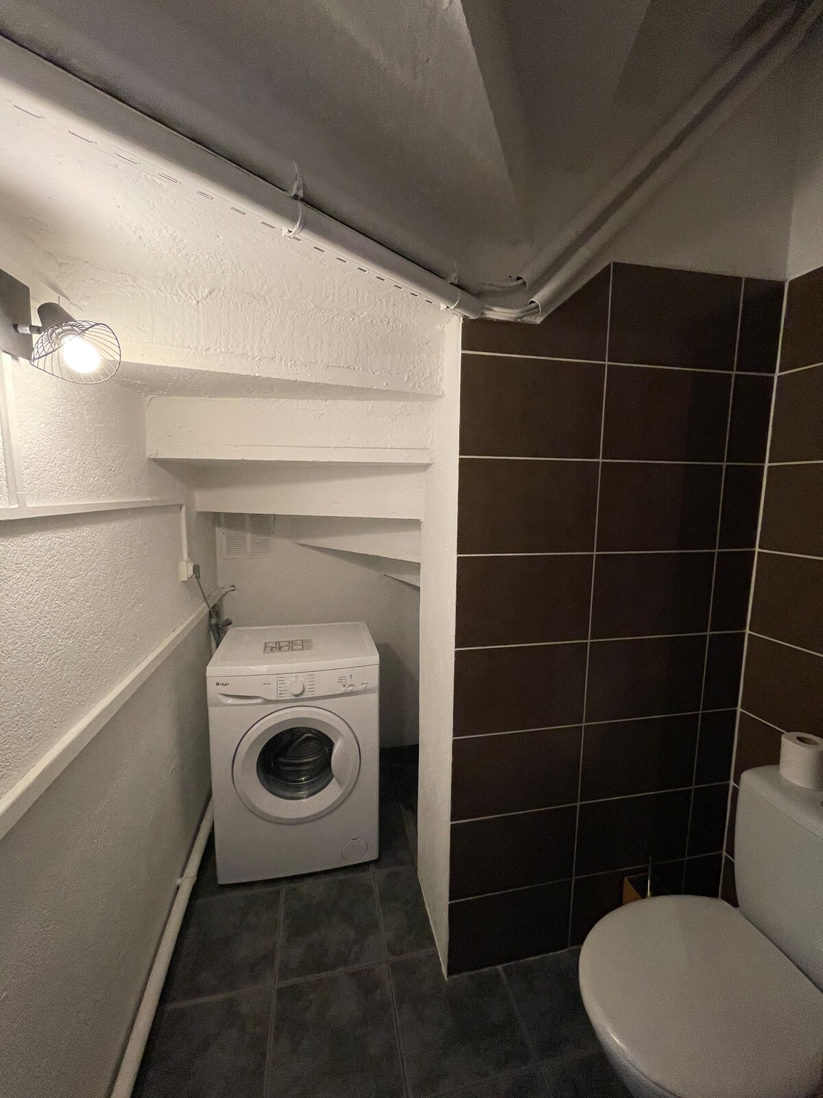 A functional laundry area is presented, featuring a white washing machine positioned beside a compact toilet. The space showcases a mix of grey and brown tiles, complemented by illuminated wall fixtures that enhance visibility.