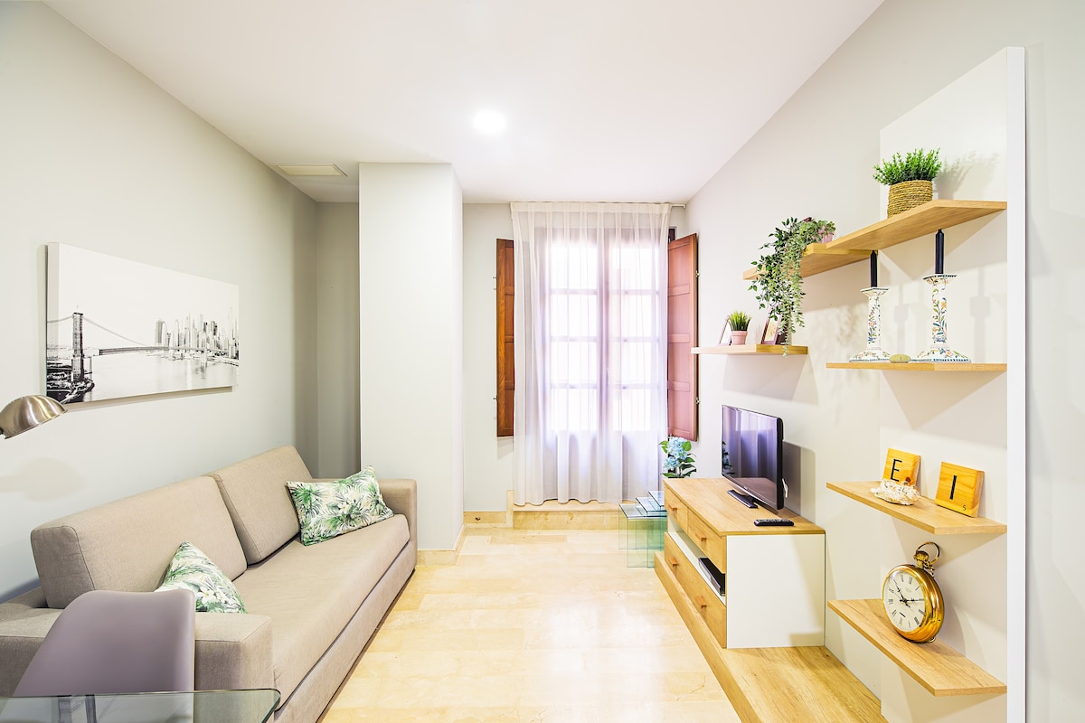 A modern living space is shown, featuring a light-colored sofa adorned with green cushions. Natural light filters through the window, complemented by wooden shutters. A TV stand with decor and a clock is present, along with a minimalist shelf displaying plants and decorative items.