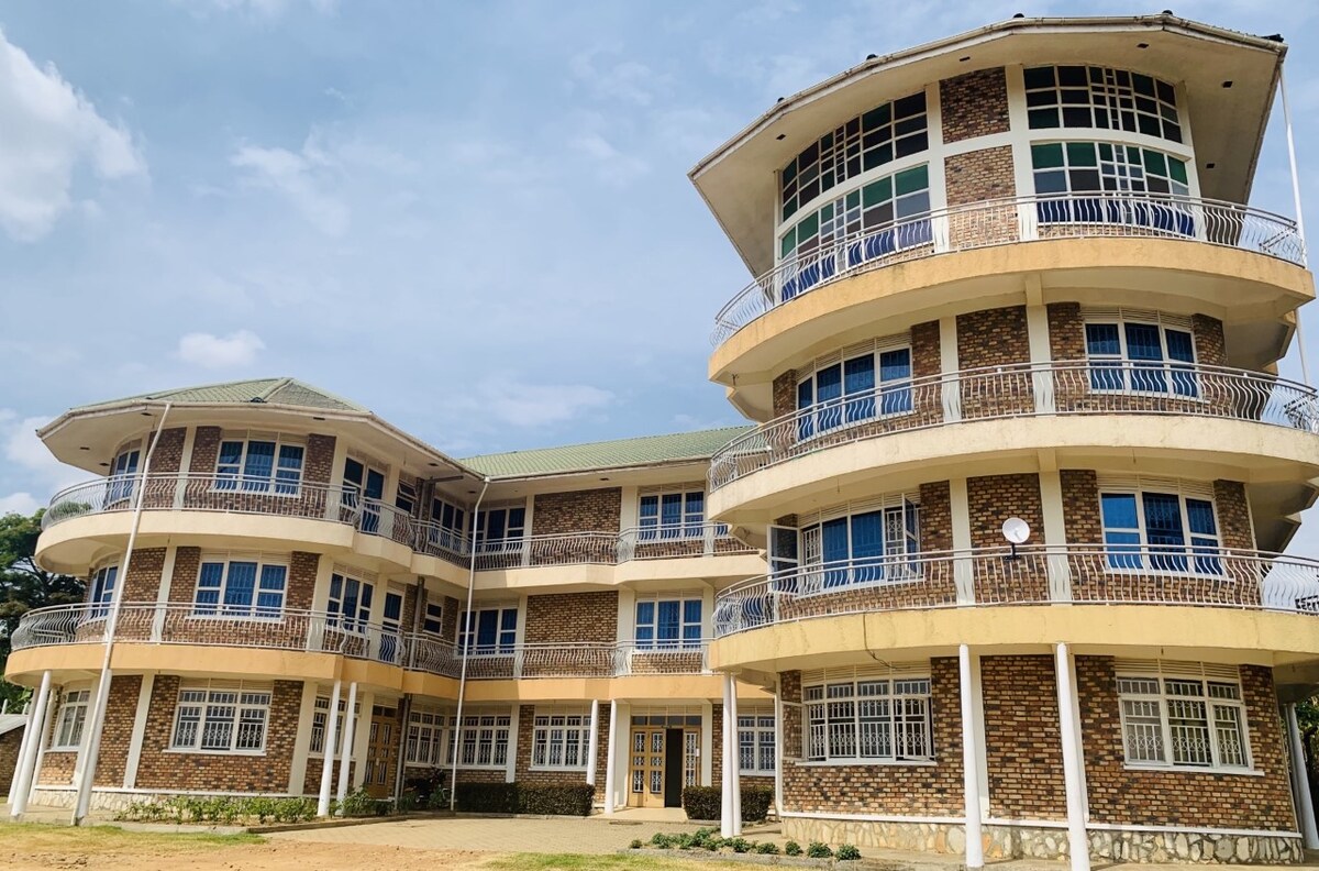 A multi-level building features a unique architecture with curved balconies, large windows, and a stone facade. The structure is surrounded by a grassy area, providing a welcoming entryway. Soft clouds can be seen in the blue sky above, complementing the sunlit exterior.