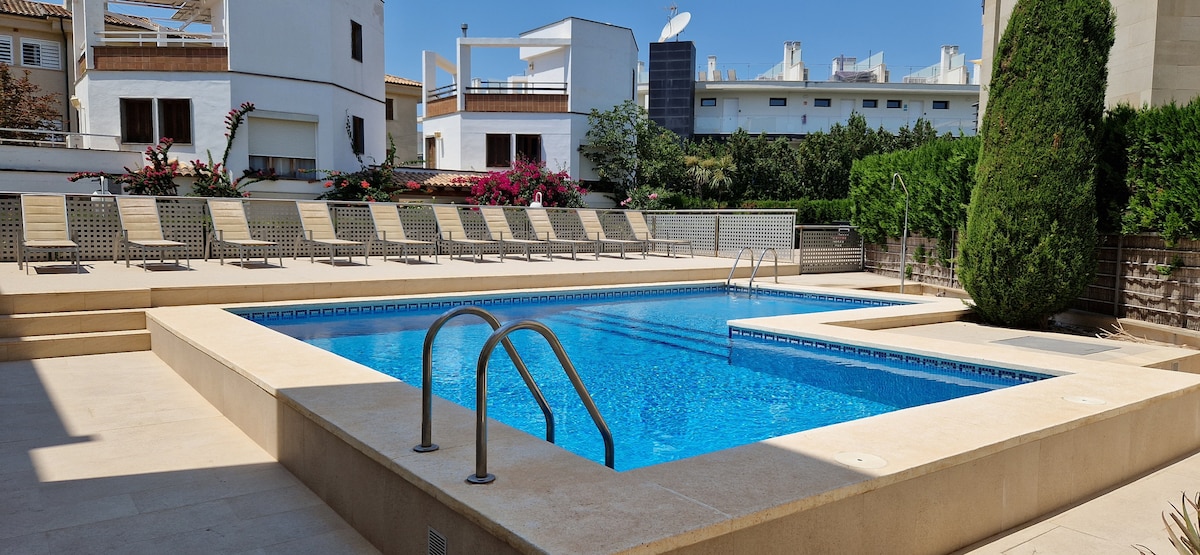 A clear view of a swimming pool is presented, surrounded by lounge chairs arranged neatly along the paved area. Lush greenery and flowering plants frame the space, contributing to a serene environment. The bright blue water reflects the sunlight under a clear sky.