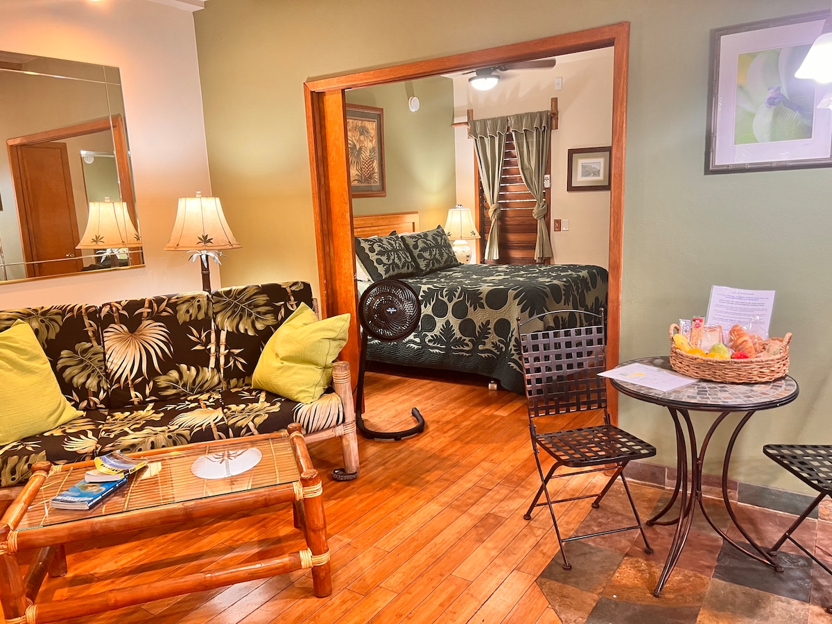A cozy living area features a tropical-themed sofa and a small dining table with two chairs. A vibrant bed with dark green bedding is visible through the open doorway. Warm wooden flooring and soothing green walls enhance the inviting atmosphere.