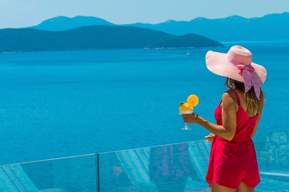 A person in a pink hat and bright red outfit stands on a terrace, holding a refreshing drink. The vast blue sea stretches out in front, with distant islands visible in the background under a clear sky, creating a serene coastal atmosphere.