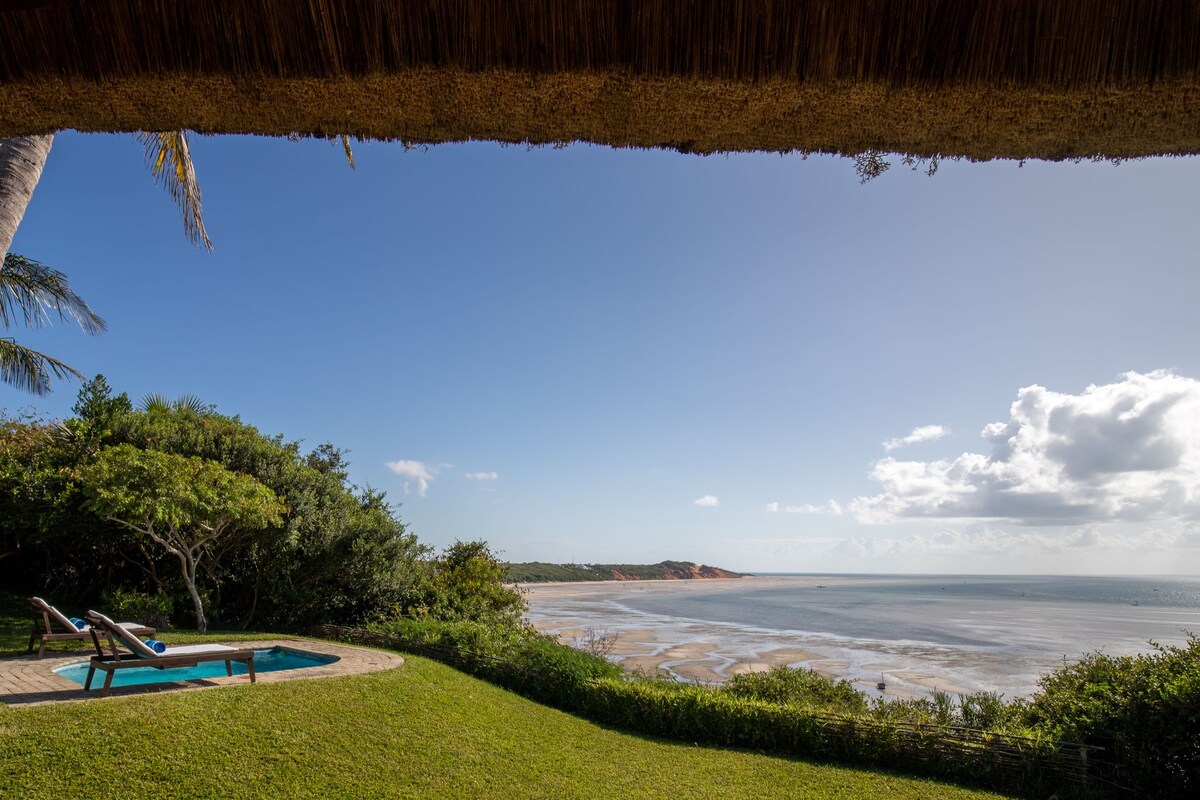 A tranquil ocean view is presented, featuring a small pool surrounded by lush greenery and a sunbed. Open skies and gentle clouds complement the seaside landscape, while distant red cliffs are visible along the shoreline.
