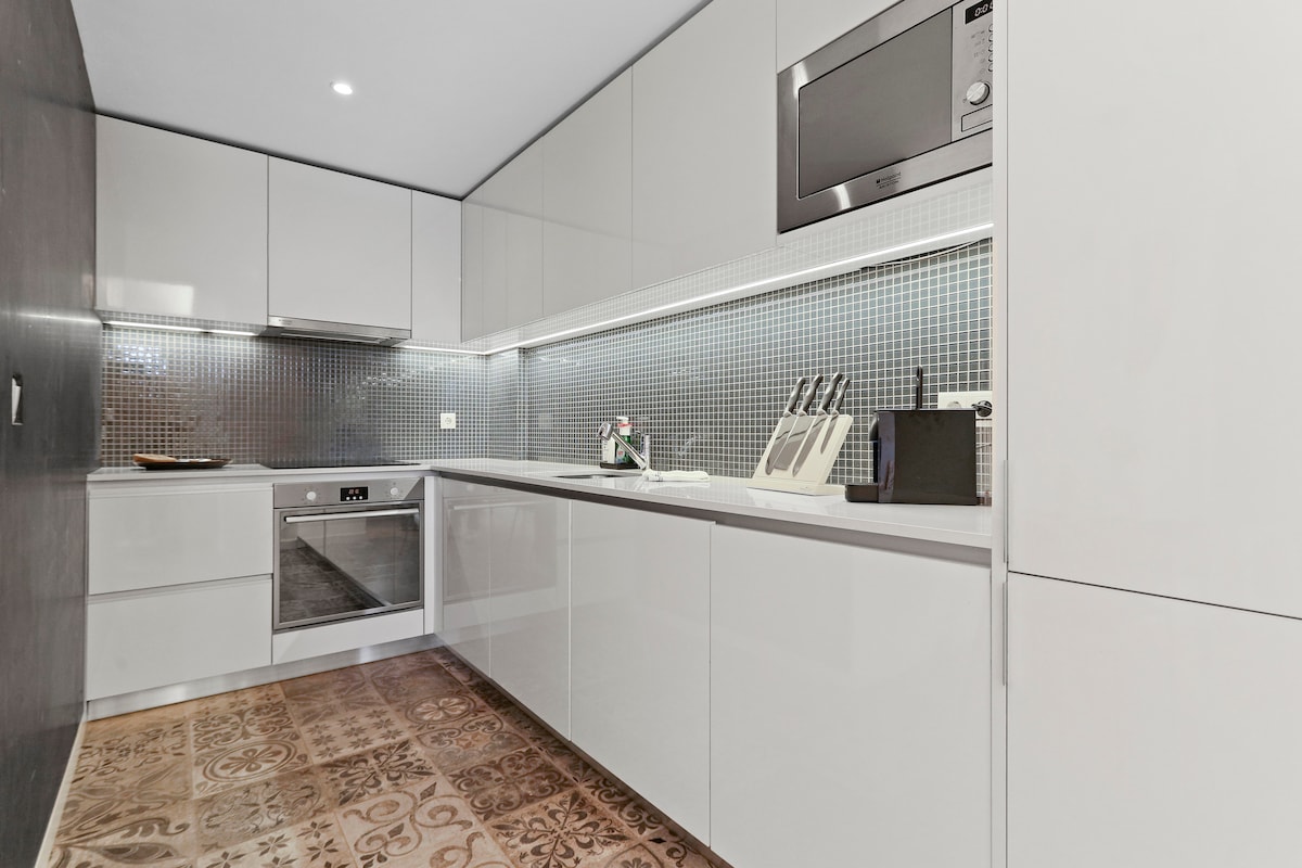A modern kitchen is presented with sleek white cabinetry and a full range of appliances. A mosaic tile backsplash adds texture. A knife block and cooking utensils are neatly arranged on the countertop, complemented by a microwave mounted above the stove.