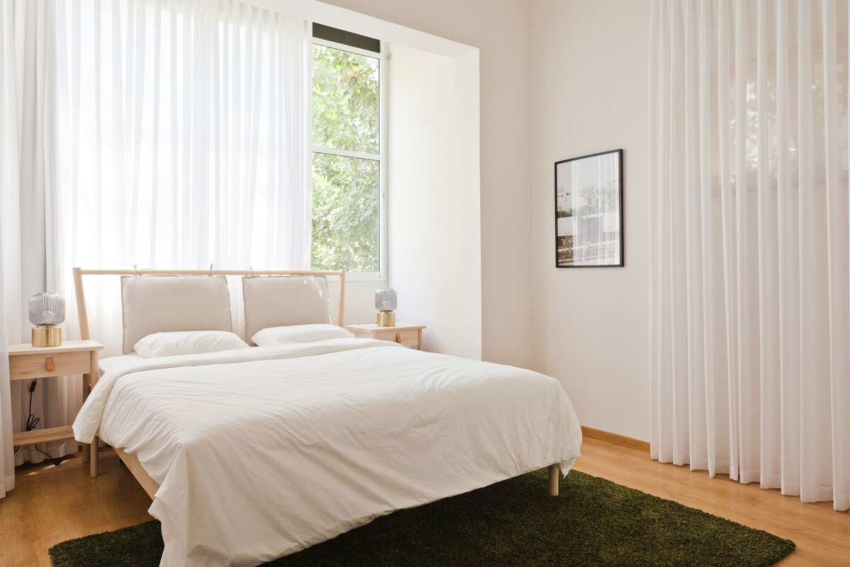 A bright bedroom features a spacious bed with a white duvet and two soft pillows, framed by large windows covered with flowing sheer curtains. Natural light fills the room, highlighting a green area rug and minimalist bedside tables with lamps, creating a calming atmosphere.