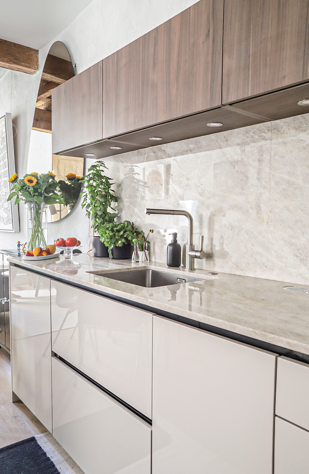 A modern kitchen space features sleek cabinetry with a combination of white and wood finishes. A marble countertop is complemented by a sink and stylish kitchen accessories. Fresh plants are arranged along the wall, adding a touch of greenery to the area.