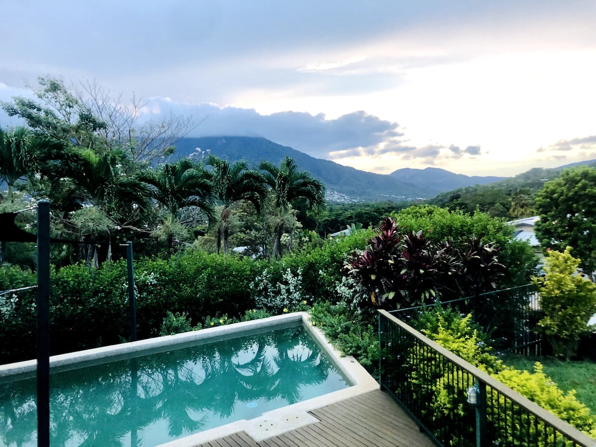 A serene view of the large pool framed by lush greenery, with mountains in the background. The sky transitions from blue to soft shades of grey and orange as the sun sets, casting gentle reflections on the water surface.