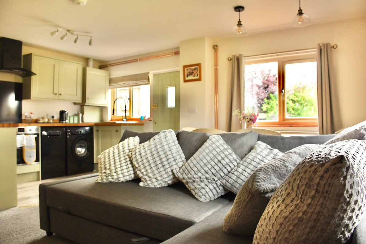 A spacious living area is presented with a comfortable gray sectional sofa adorned with decorative pillows. Natural light enters through a nearby window, complementing the neutral color palette of the space. A well-equipped kitchen is visible in the background, featuring modern appliances.