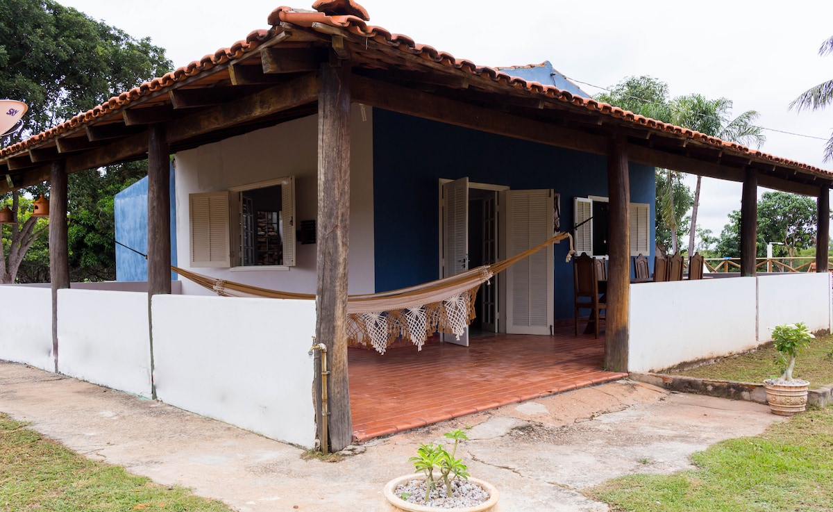 A charming exterior view showcases a single-story building with a thatched roof. A hammock is hung on a spacious porch, bordered by white walls. Lush greenery surrounds the area, enhancing the inviting outdoor setting. Large windows allow natural light into the interior.
