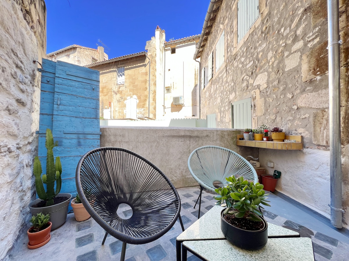 A courtyard terrace is equipped with two modern chairs and a small table. Various potted plants add greenery, while a blue wall segment provides a vibrant backdrop. Stone walls frame the area, blending the modern decor with the historical architecture.