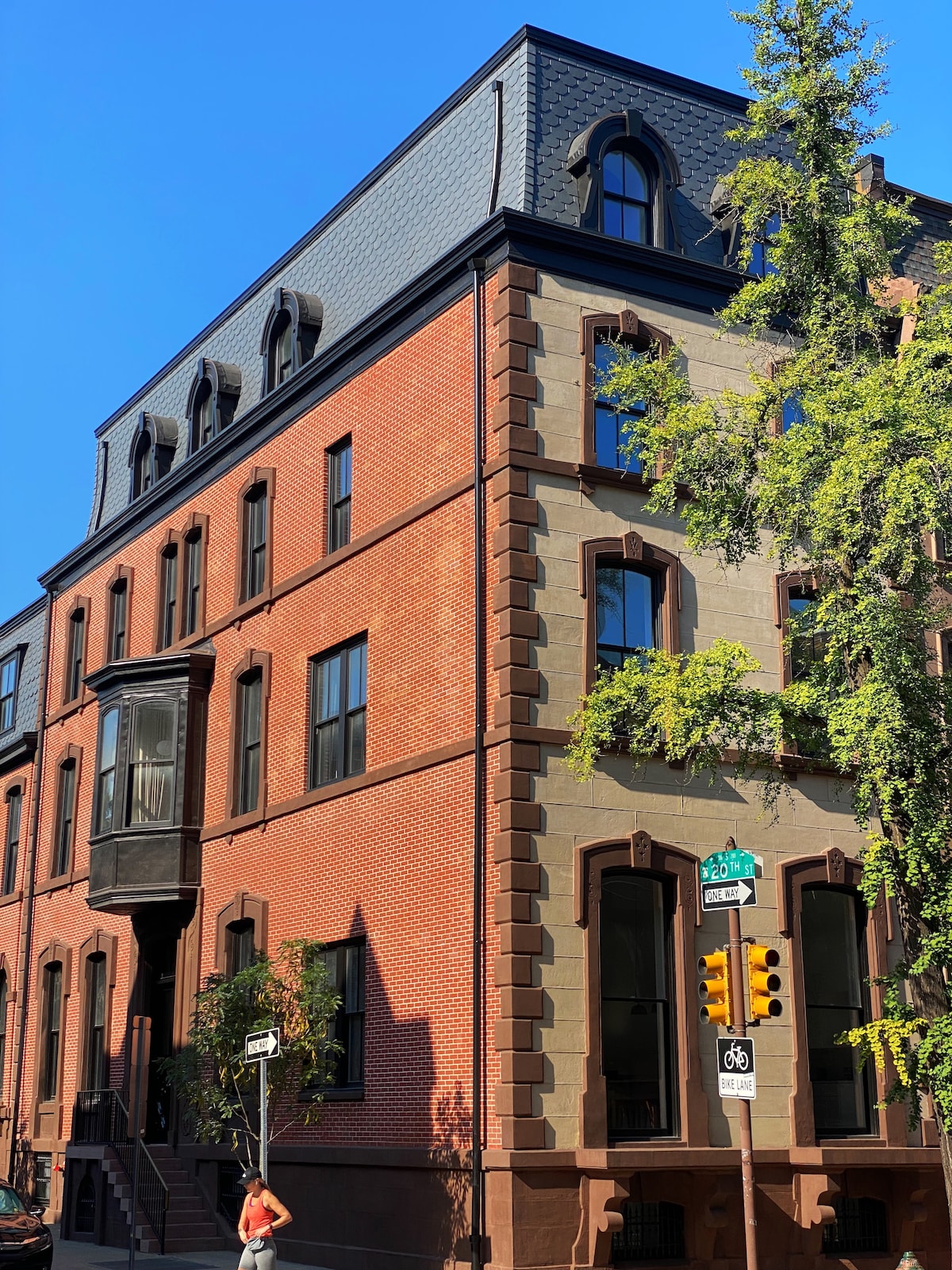 A historic building with an elegant blend of red brick and stone exteriors stands at the corner of a sunny street. The architecture showcases large windows and a prominent entrance, enhancing the character of the neighborhood.