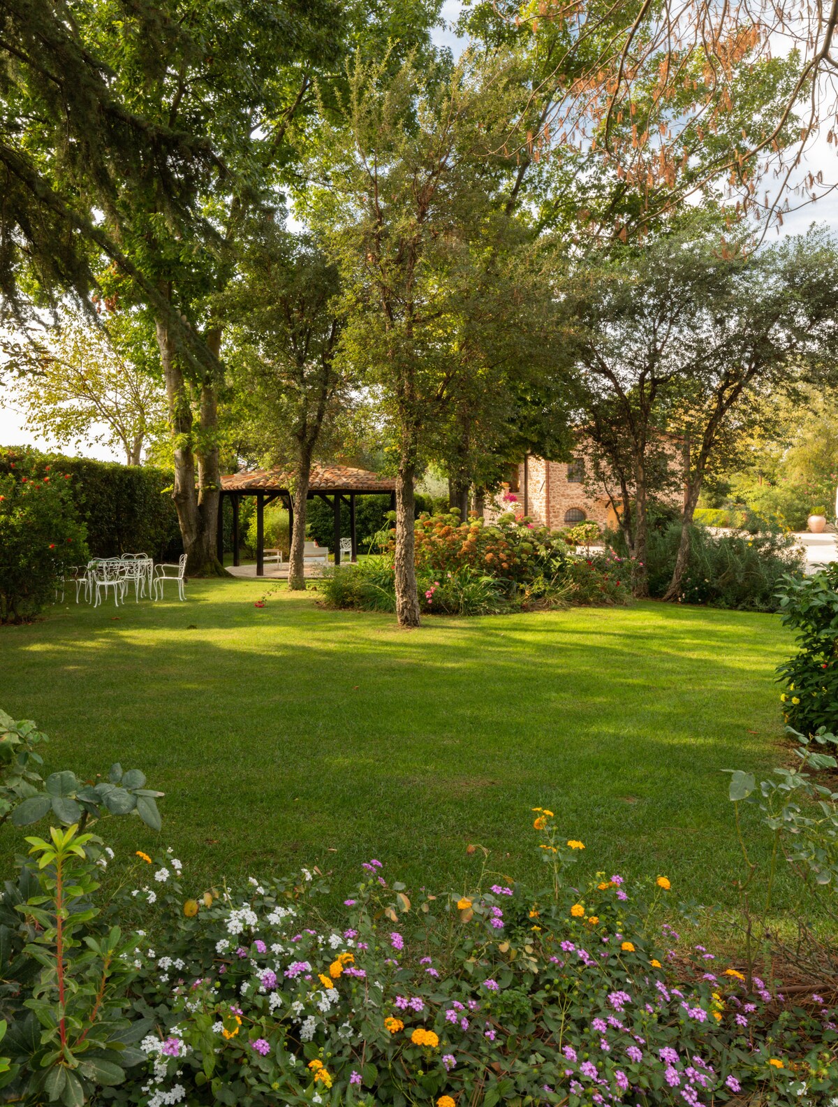 A garden area is depicted, featuring lush green grass and a variety of flowering plants. Several white chairs are arranged under a wooden gazebo, surrounded by trees and shrubs. The background includes a stone building, adding character to the serene outdoor setting.