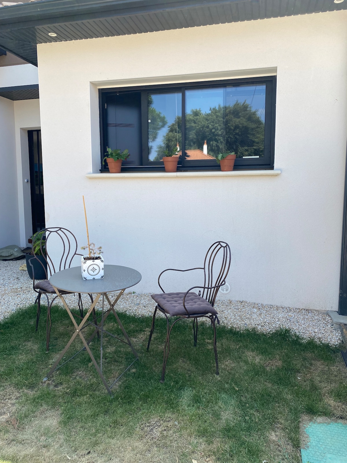 A small outdoor seating area is presented, featuring a round metal table surrounded by two decorative chairs. Potted plants are placed on the windowsill, complementing the neutral exterior wall. The grassy ground adds a natural touch to the outdoor space.