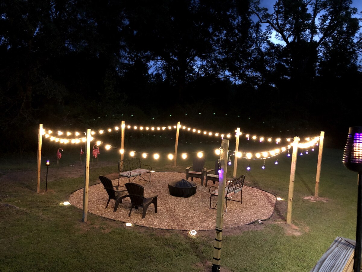 A serene outdoor gathering area is illuminated by string lights, creating a warm ambiance. Several comfortable seating options, including chairs and a fire pit, are arranged on a gravel surface surrounded by grass. Lush trees form a natural backdrop, enhancing the peaceful setting.