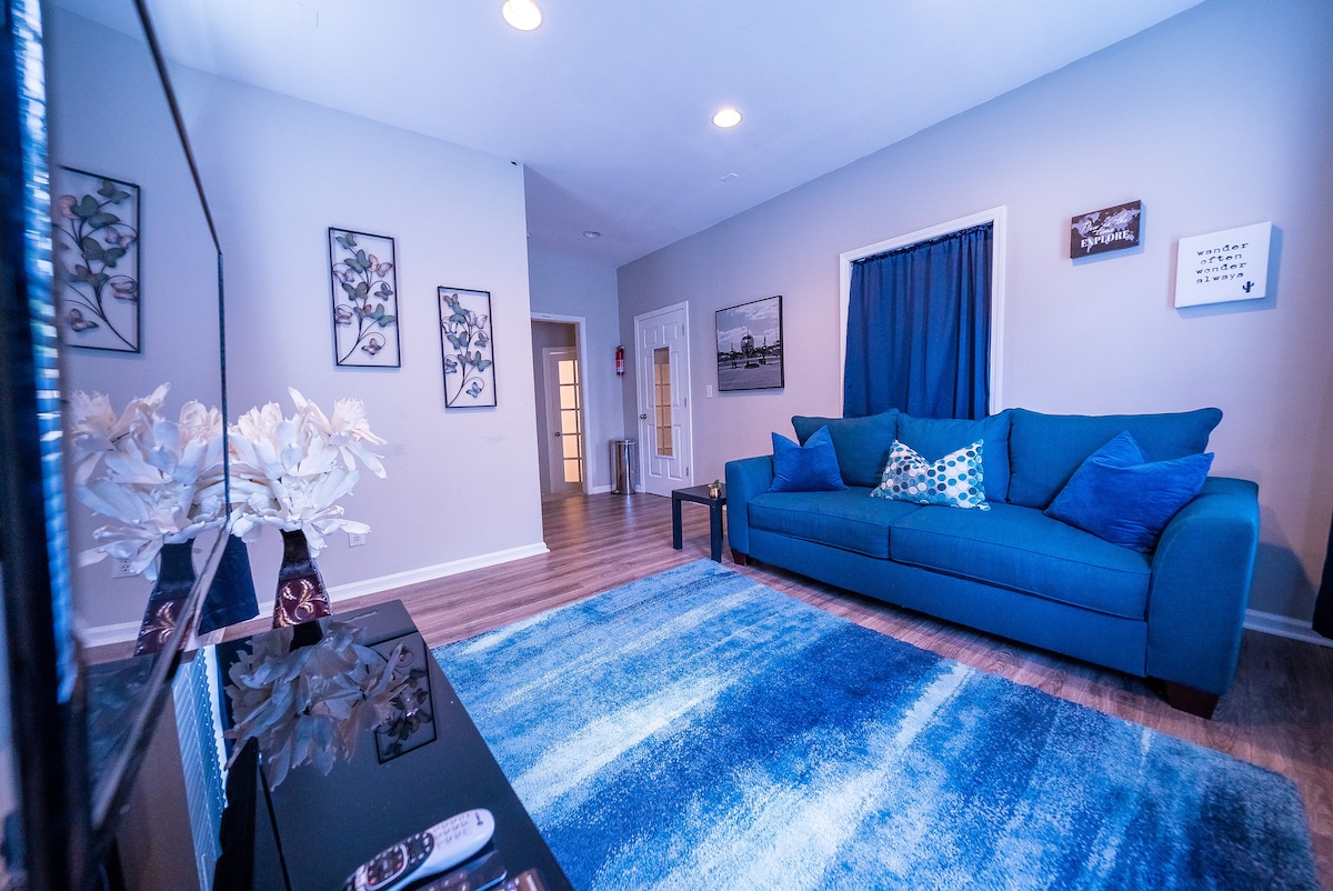 A comfortable living area showcases a vibrant blue sofa adorned with patterned cushions. A matching area rug lies beneath, enhancing the space. The room features wall art with floral themes, smooth flooring, and a television console. A curtain covers a nearby window, allowing soft light into the room.