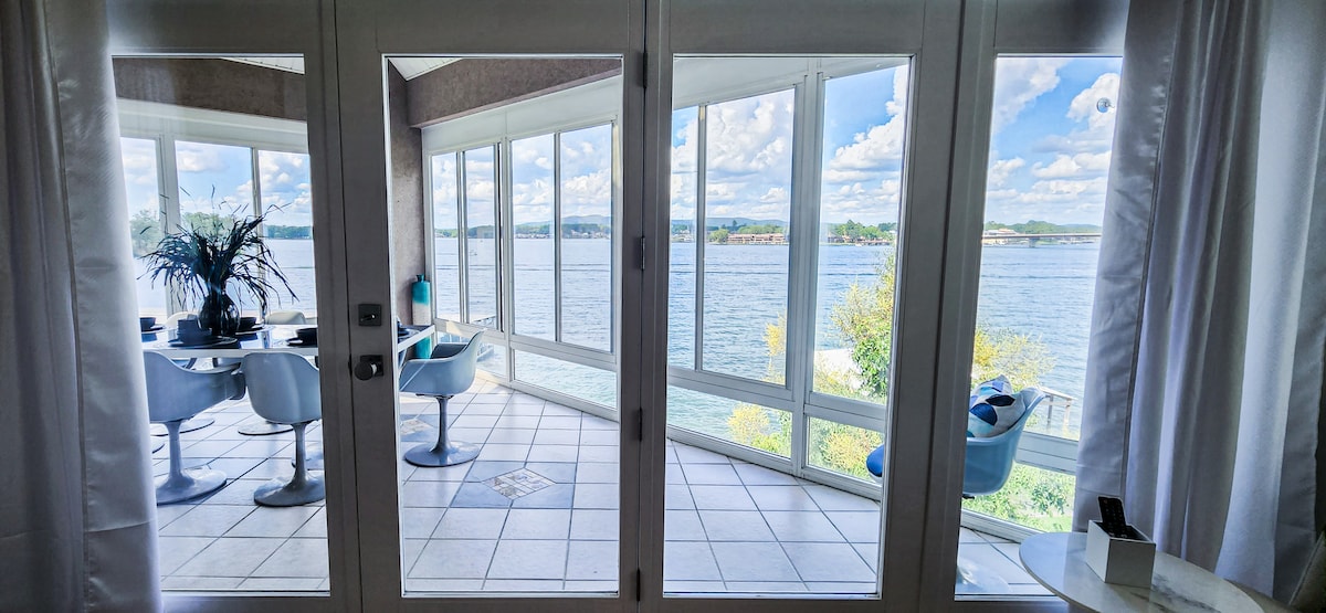 Floor-to-ceiling windows frame a stunning view of Lake Hamilton, allowing natural light to fill the space. The room features modern furniture, including a dining table and chairs, set against the tranquil backdrop of the water and skies.
