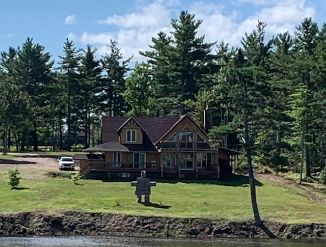 A riverside cabin is seen surrounded by tall trees, with a spacious lawn leading up to the water's edge. The structure features a large porch and multiple gabled roofs, all under a clear blue sky. A parked car is visible near the cabin.