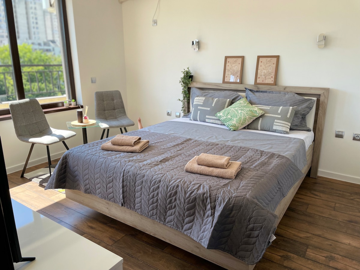 A spacious bed with a textured gray quilt lies at the center of the room. Two gray chairs are positioned alongside a small table. Natural light fills the space through a large window, enhancing the minimalist decor showcased by framed art on the walls.