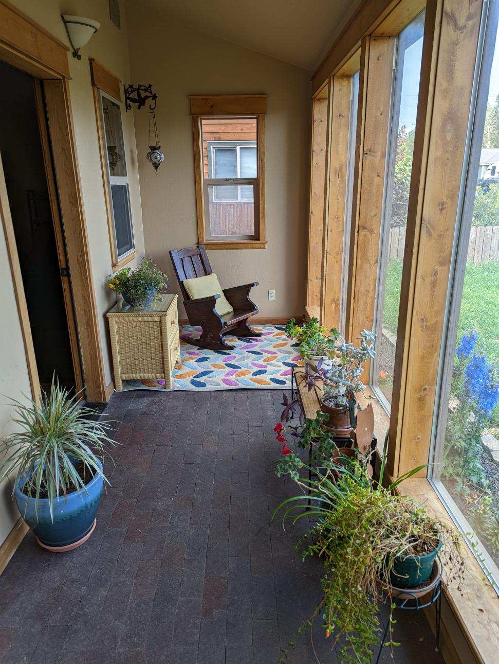 A sunlit sunroom with large windows showcasing greenery outside. A wooden rocking chair is positioned beside a small side table, adorned with potted plants. A colorful rug adds texture to the floor, while additional plants are displayed in various pots along the space.