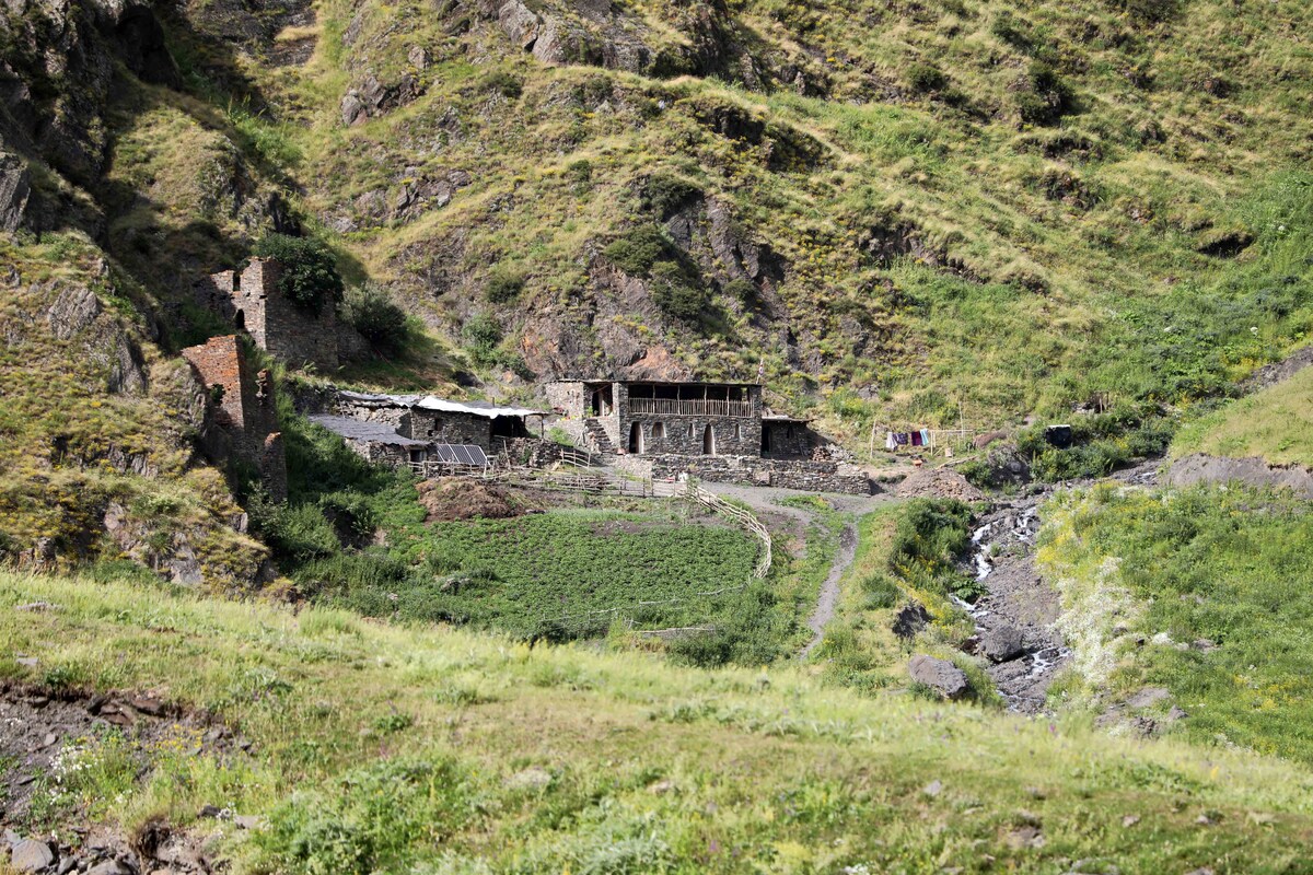 A remote landscape showcases an abandoned stone structure nestled among green hills. A small stream winds through the area, while remnants of past settlements are visible, surrounded by lush vegetation and rocky terrain, invoking a sense of untouched nature.