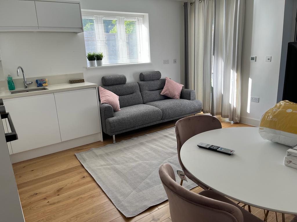 A bright living space is presented, featuring a gray sofa with pink cushions and a round dining table surrounded by four chairs. Light enters through a window, while drapes frame the view. A rug lies on the wooden floor, adding warmth to the area.