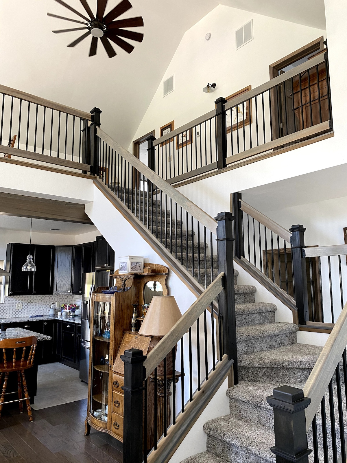 A well-lit staircase with a mix of light and dark wood features rises to a second floor. The railing is designed with black spindles, and a spacious open-air feel is created by high ceilings. A glimpse of the kitchen can be seen on the lower level.
