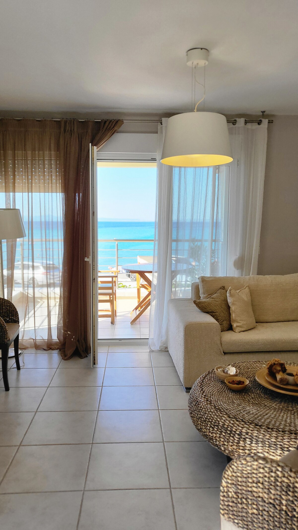 The living area is filled with natural light, featuring a light-colored sofa and a round coffee table with refreshments. Large glass doors open to a balcony overlooking the sea, while sheer curtains provide a soft separation from the outdoors.
