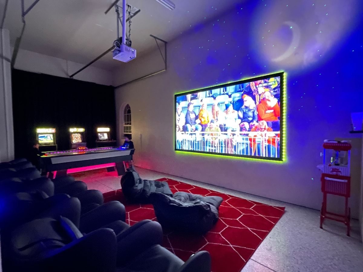 An entertainment area is showcased with a large HD projector screen displaying a lively scene. Comfortable seating options, including massage chairs and bean bags, are arranged in front. Two arcade machines add a touch of fun, framed by soft lighting that enhances the atmosphere.
