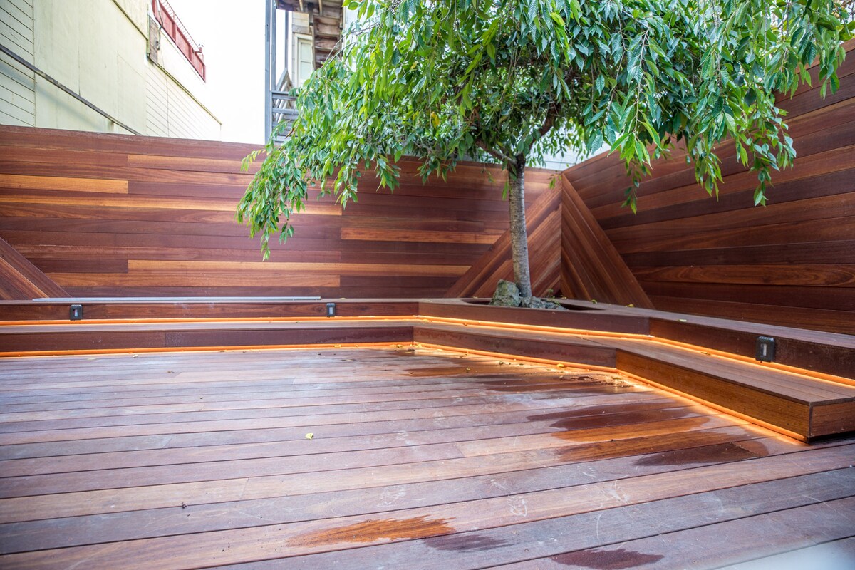 A spacious backyard deck features warm wooden tones with built-in seating areas illuminated by soft lighting. A healthy tree stands at the center, complemented by the surrounding wooden paneling, creating a serene outdoor environment.