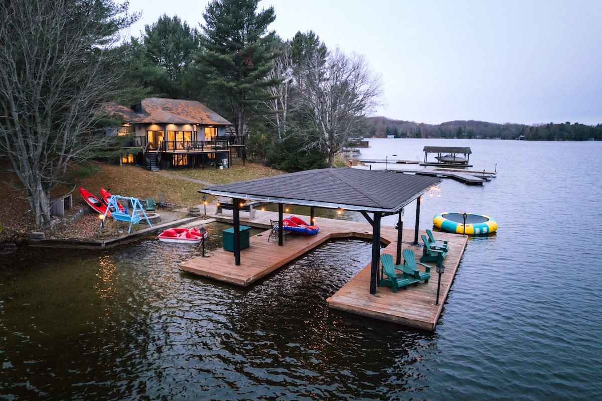 A serene waterfront area features a wooden dock with ample seating and a designated space for watercraft. Nearby, kayaks, a paddle boat, and a colorful inflatable banana boat are arranged on the shore, with lush greenery and calm water surrounding the space.