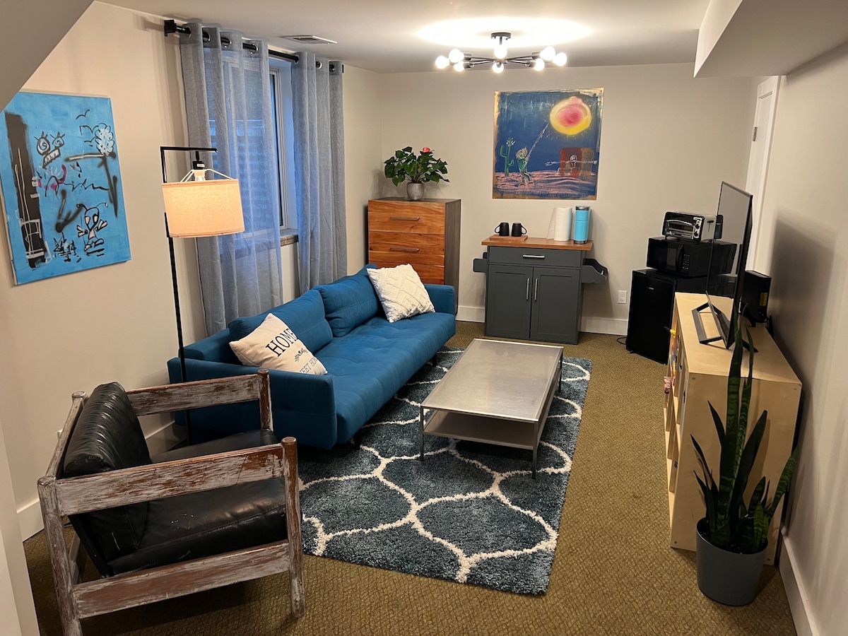 The living area features a blue sectional sofa and a black chair positioned around a low coffee table, with a textured rug underneath. A small console and a TV stand are present, accompanied by a colorful wall art piece. Natural light filters through sheer curtains.