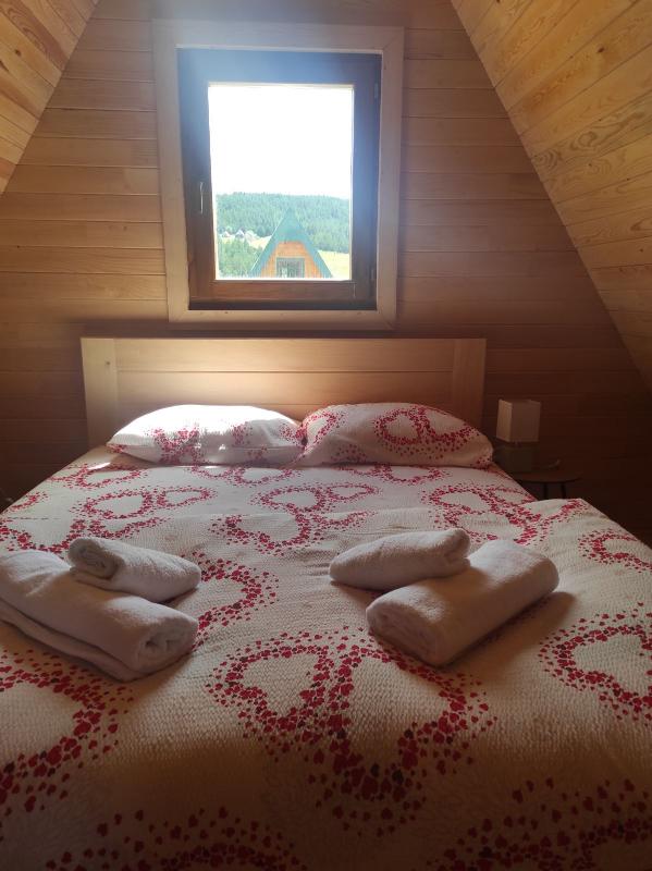 A warmly designed sleeping area features a spacious bed adorned with a patterned blanket and colorful pillows. Soft towels are neatly arranged on the bed, while a window allows natural light to brighten the wooden walls and offers views of the outdoors.