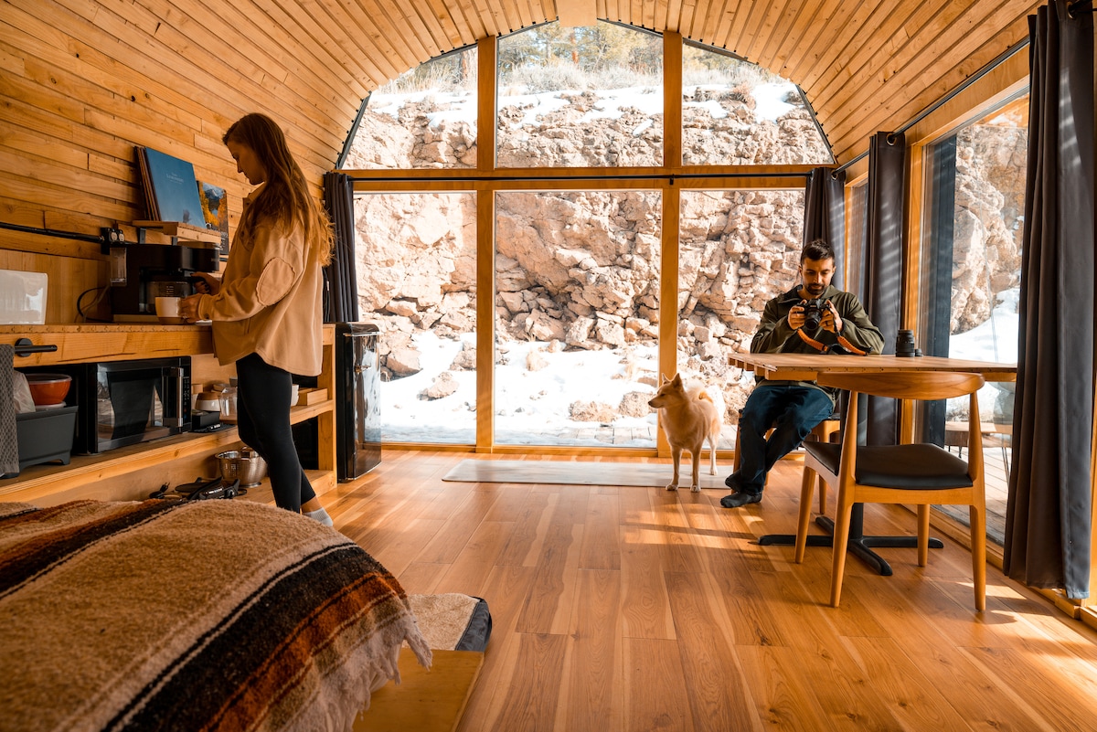 The interior of the Rock Room features warm wooden walls and flooring, showcasing a cozy bed adorned with a patterned blanket. A seating area with a table is visible, alongside a kitchenette with essential amenities. Large windows offer a view of a rock wall and natural surroundings.