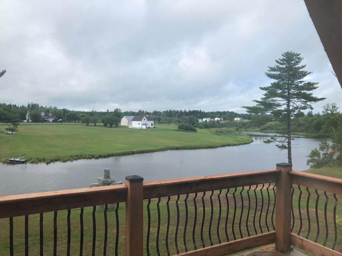 A scenic view of the Tracadie River is captured from the balcony, showcasing lush green landscapes on either side. A small boat is visible on the water, while a few houses can be seen in the distance, framed by trees and gentle clouds in the sky.