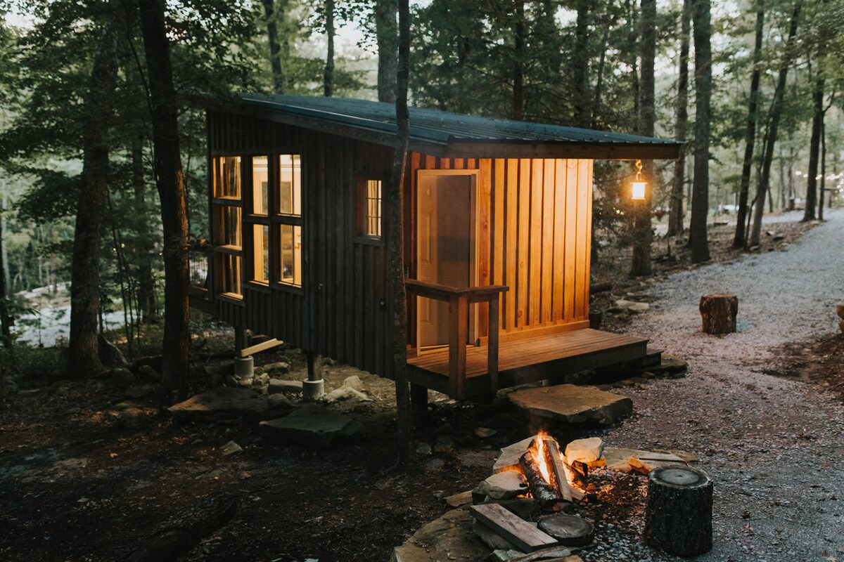 An elevated cabin crafted from wood is nestled among trees, illuminated by soft lighting. A small porch is accessible through a wooden door, while a fire pit rests nearby on a stone surface, surrounded by logs for seating.