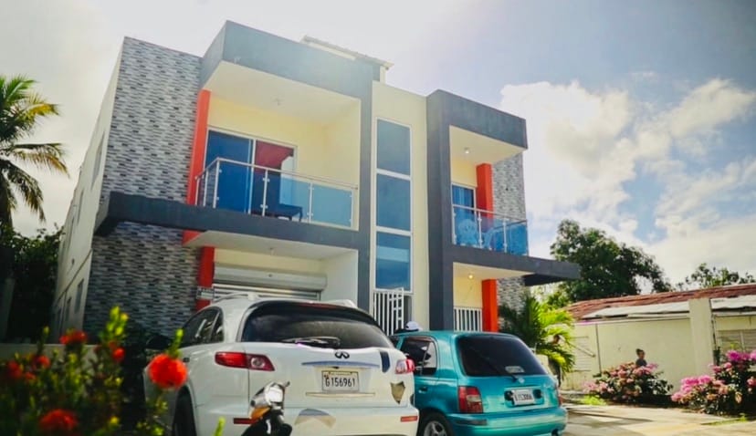 The modern building features two distinct levels with large glass balconies, allowing natural light to fill the interior. A white car and a light blue vehicle are parked in front, surrounded by lush greenery and colorful flowering plants, enhancing the vibrant atmosphere.